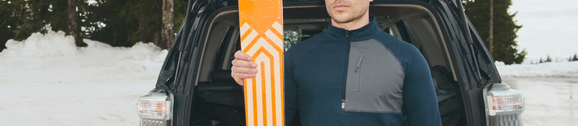 Man outside in snow wearing heavyweight baselayer holding skis beside vehicle