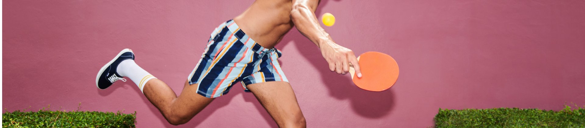 Man in swim shorts against pink wall playing ping pong