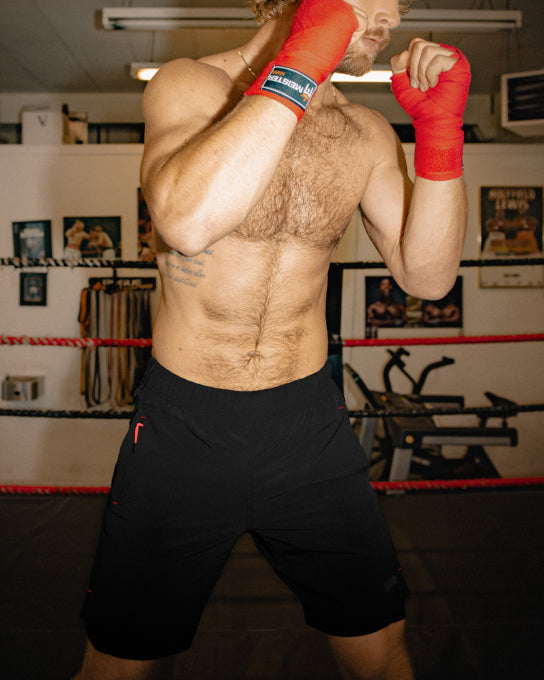 A man in a boxing ring with his fists raised to his face. His hands are wrapped in red boxing tape. He wears black shorts.
