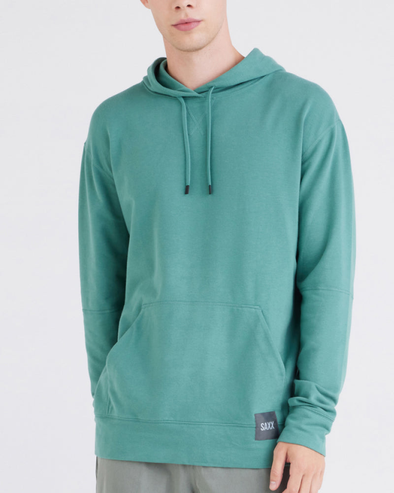 A close up of a man wearing a turquoise hoodie with a black box logo at the the left-hand side hem.