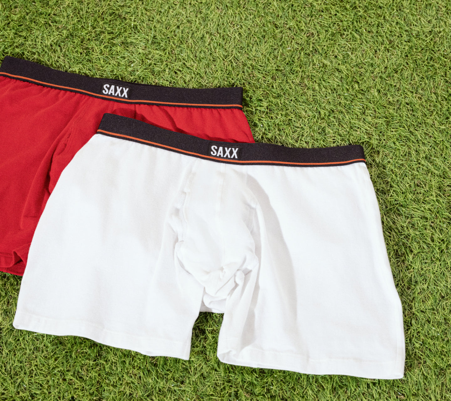 Two pair of boxer briefs, one white and one red laid on astro turf. The white pair is laid slightly on top of the red pair.