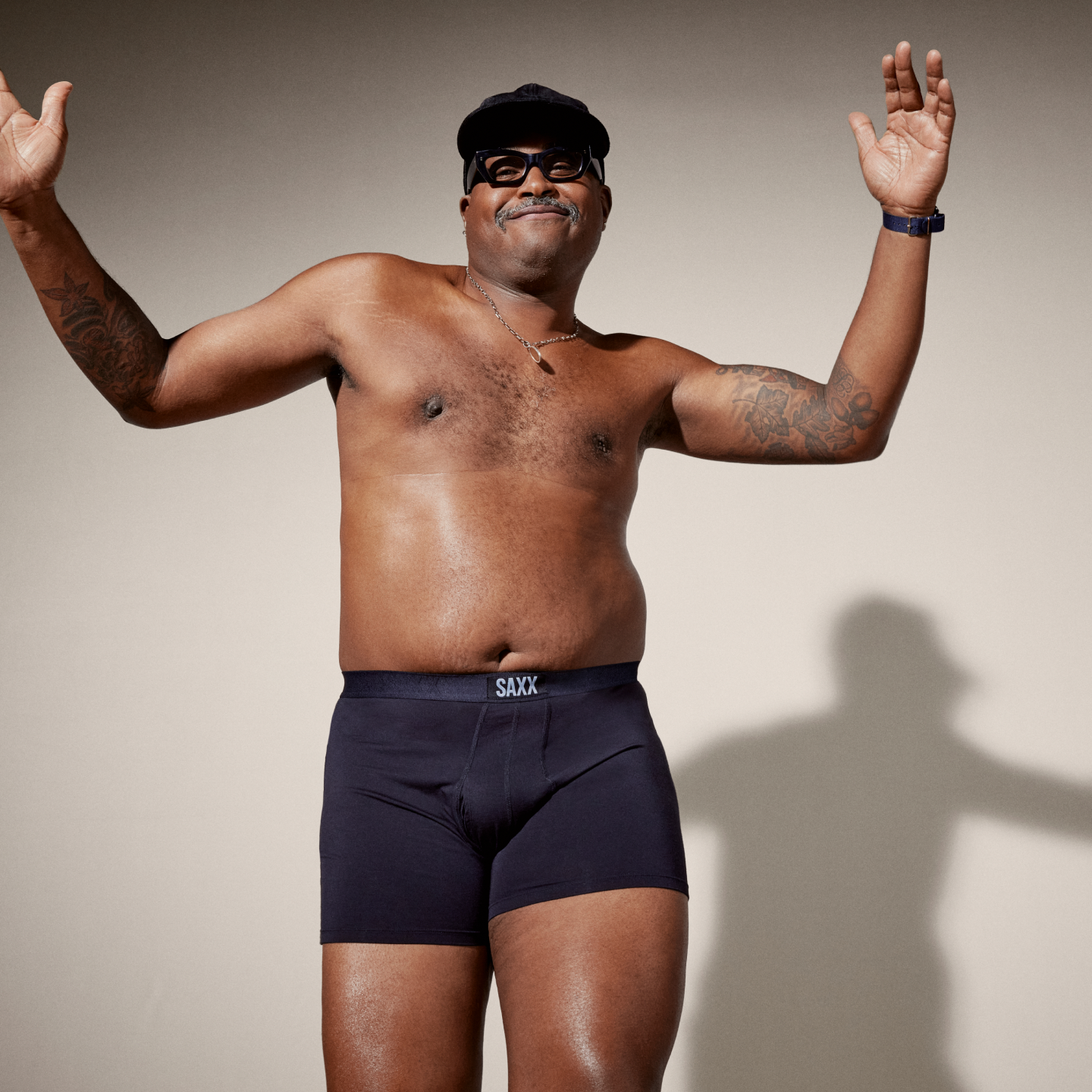 A middle-aged man dancing against a neutral backdrop wearing black SAXX boxer briefs, a necklace, black-rimmed glasses, and a black baseball cap. 