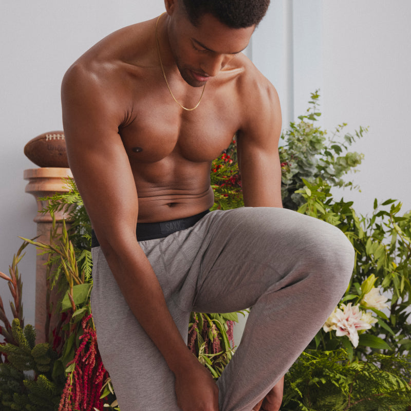 A man wearing grey SAXX Sleep Pants adjusting his ankle cuff while standing in a room filled with winter greenery. 