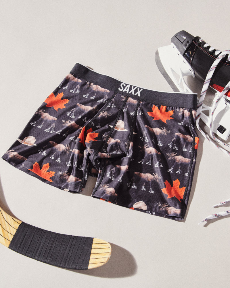 Pair of black SAXX Boxer Briefs with Canadian-themed print laid flat over neutral backdrop near hockey stick and skates.