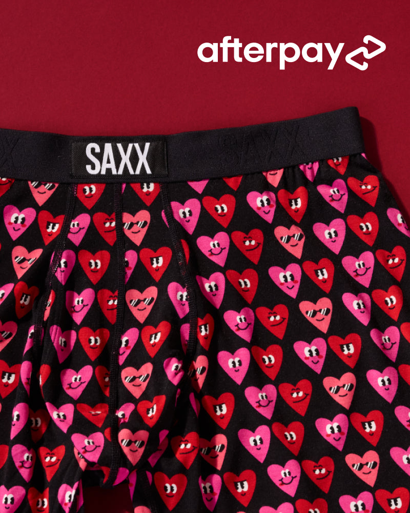 Black Valentine’s themed SAXX Boxer Briefs with pink and red smiling hearts pattern over red background. Text reads ‘Afterpay’.