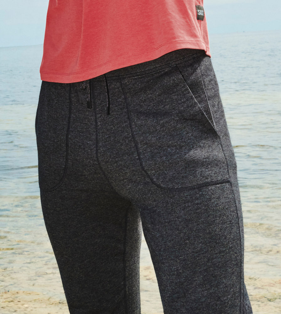 Closeup on man’s lower body on the seashore wearing dark grey heather SAXX activewear bottoms and red SAXX shirt.