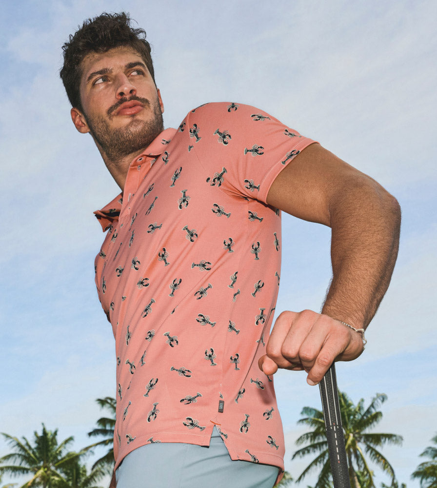 Man in salmon SAXX Polo shirt with lobster print, arm leaning on golf club, looking offscreen, palm trees behind.