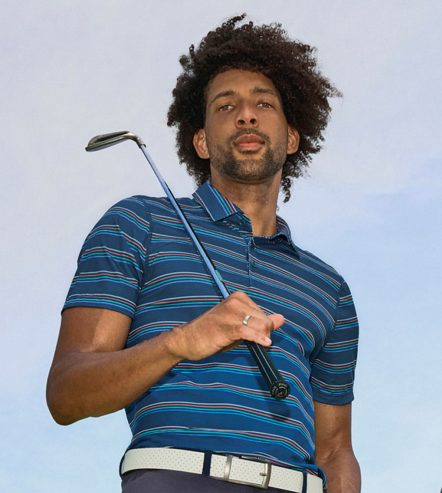 Man in blue striped SAXX Polo shirt looking direct to camera holding golf club, behind blue sky.