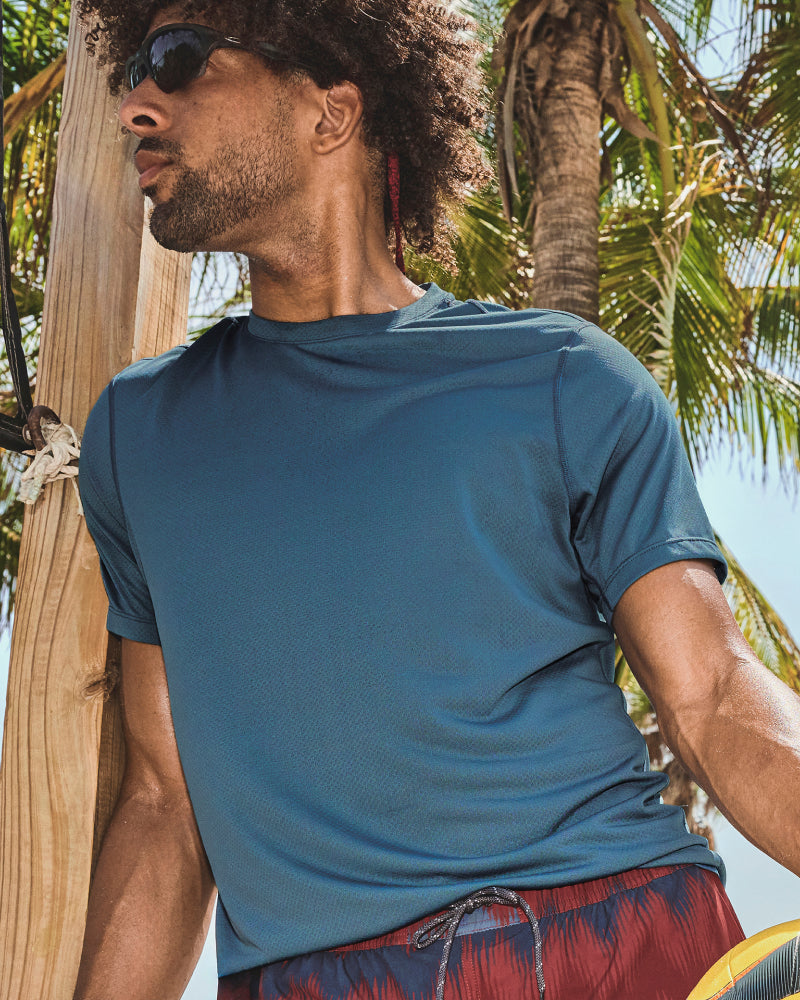 Man in blue SAXX Multi-Sport Tech Tee and red and blue SAXX 2N1 Shorts looking into the distance, palm trees behind.