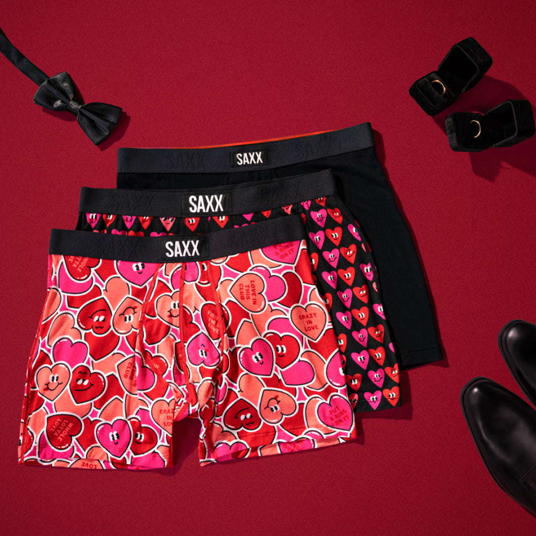 Three pairs of Valentine’s Day themed SAXX Boxer Briefs laid out flat over red backdrop, black bowtie and dress shoes nearby.