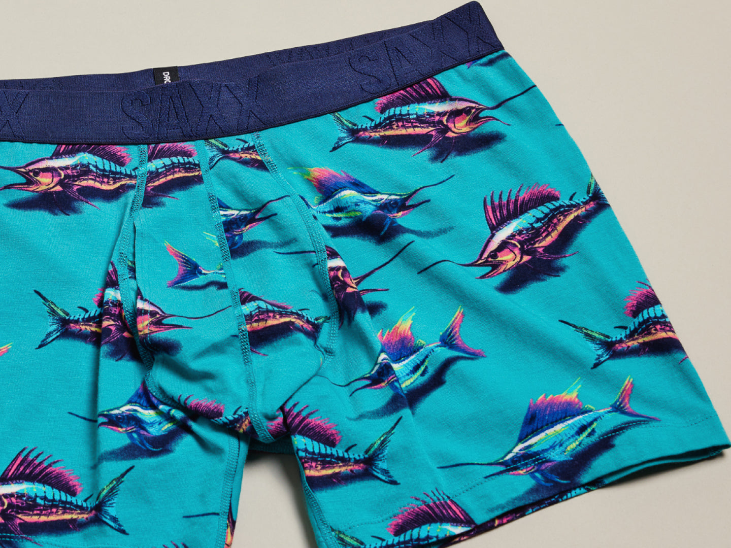 Image of green swordfish printed SAXX Trunks and a folded multi-colored pair on a neutral backdrop.