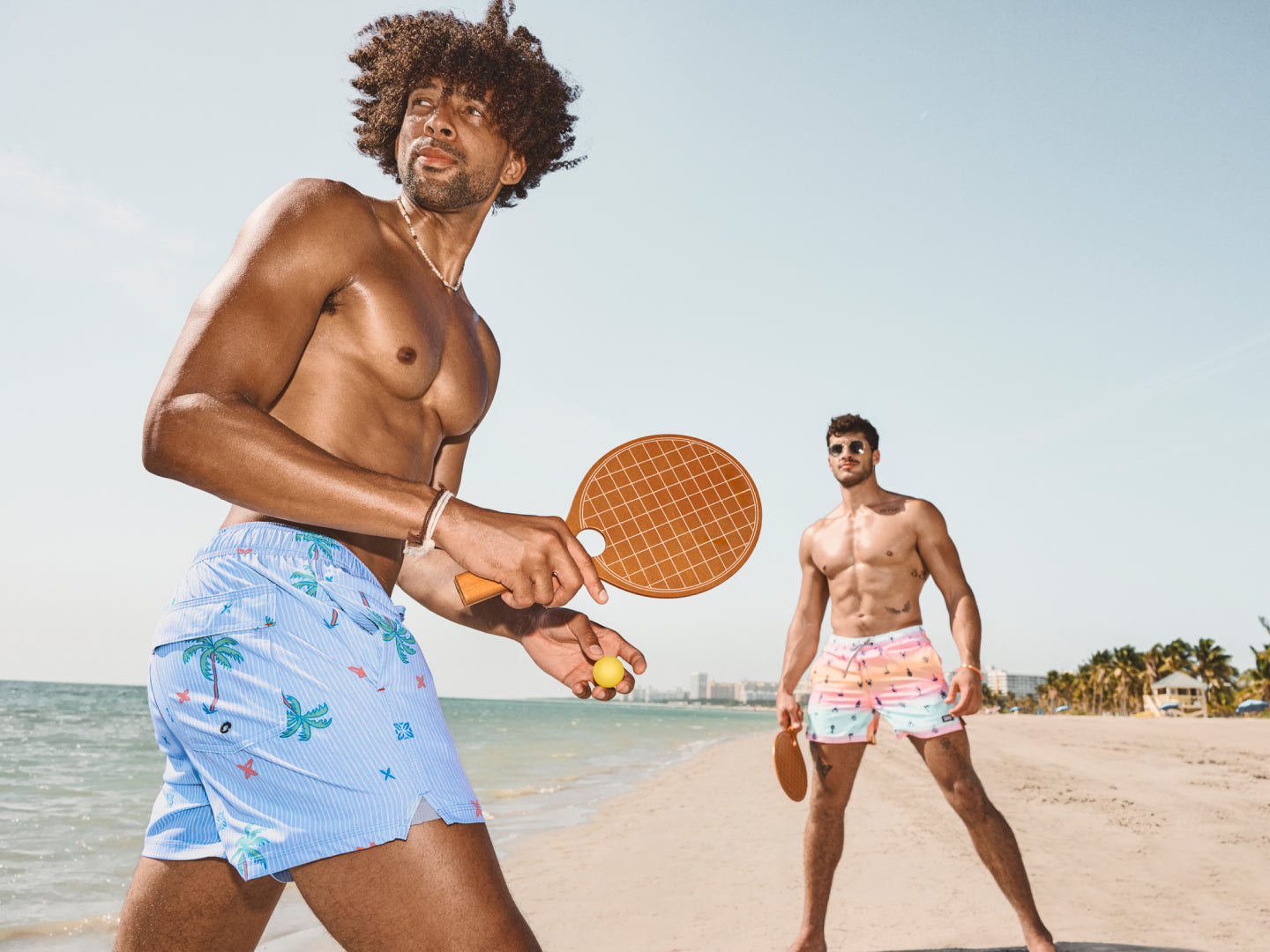 Two fit men wearing multi-color SAXX Swim Shorts on the beach playing paddle ball.