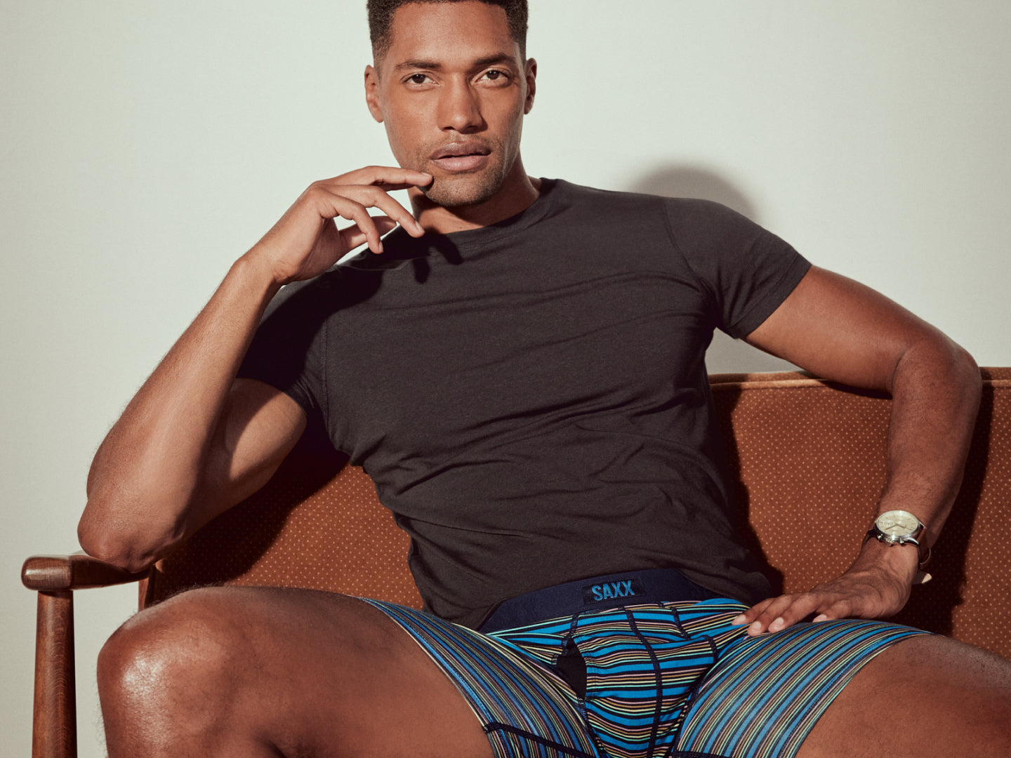 A closeup of a male model sitting on a brown couch wearing blue striped SAXX Boxer Briefs and a black SAXX tee.