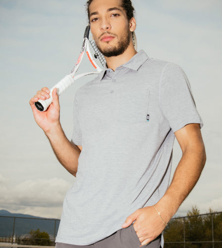 A man standing on a tennis court, mountains visible behind him, wearing a grey SAXX polo shirt and grey SAXX 2N1 Shorts holding a tennis racket. 