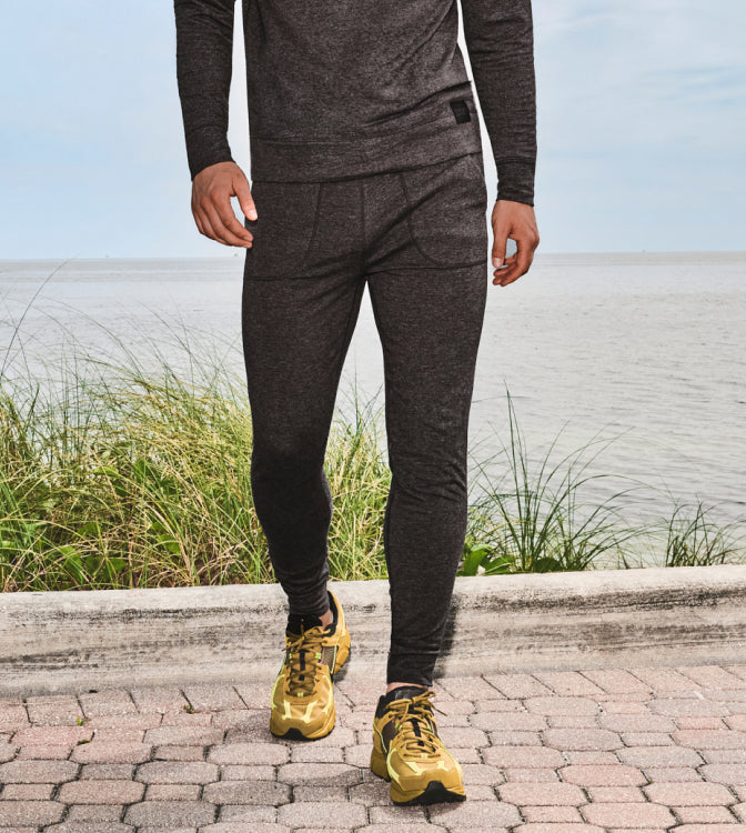 Man standing on sidewalk by ocean wearing matching dark grey set of SAXX lounge wear and gold running shoes.