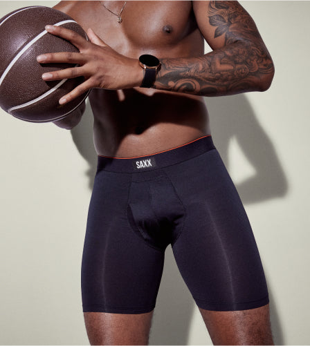 A man standing against a neutral backdrop wearing SAXX Boxer Briefs and a black watching, holding a basketball.