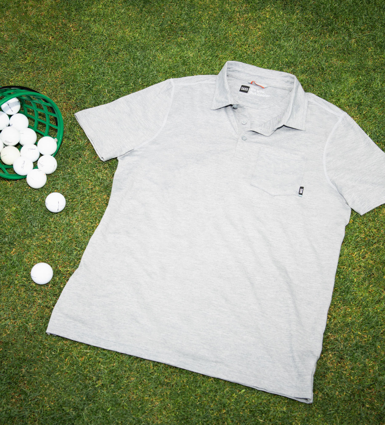 A white polo shirt laid flat, face-up on a putting green with a bucket of golf balls seen on the left of the image. 