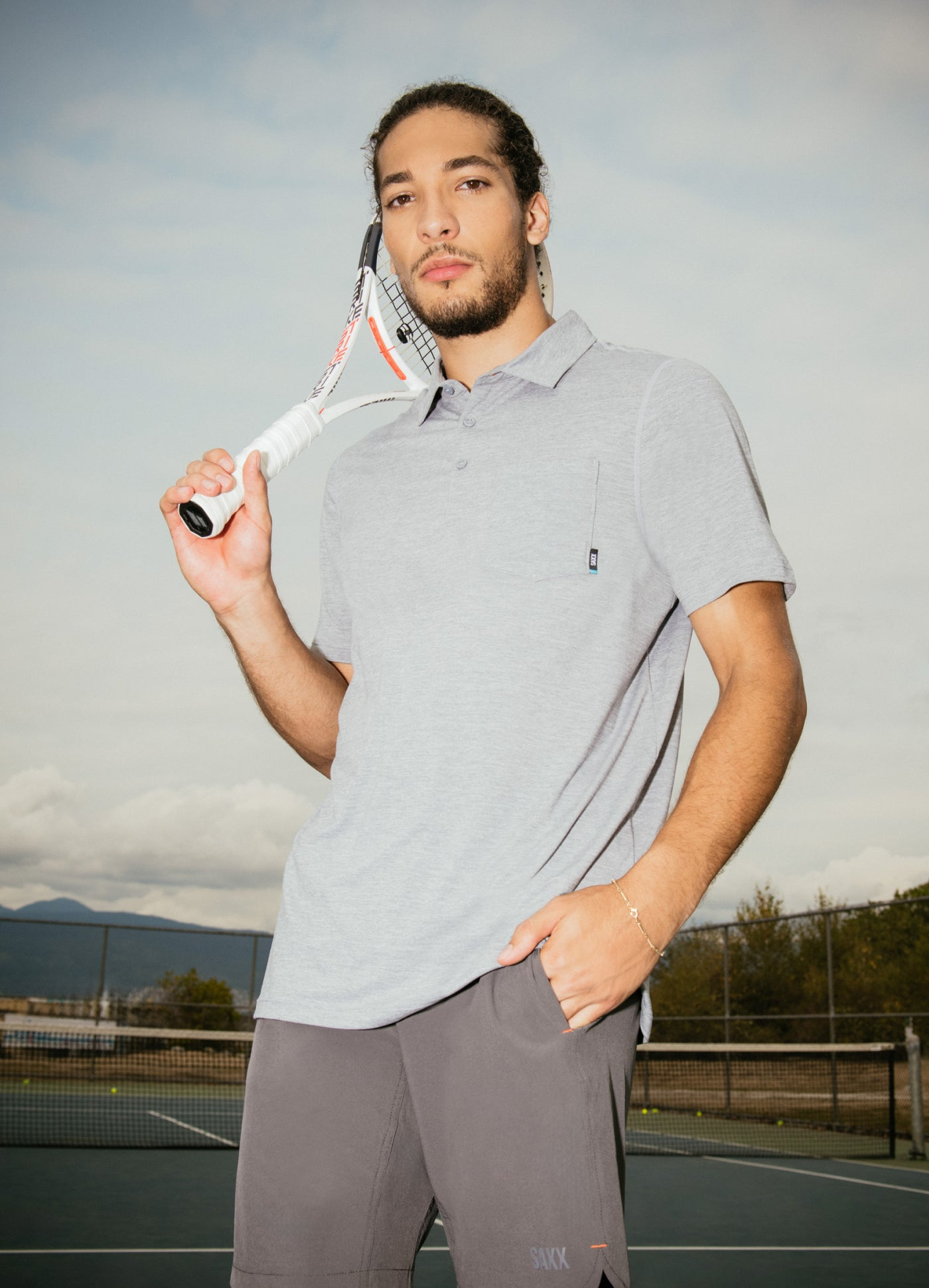 A man seen from hip up. He wears a grey polo. He holds a tennis racket in his right hand and rests it on his right shoulder.