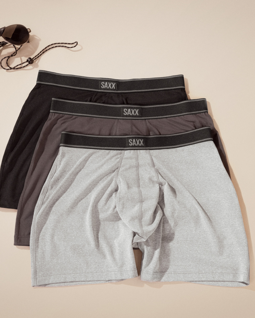 Three pairs of SAXX Boxer Briefs in light grey, dark grey, and black, laid out neatly over a neutral background.