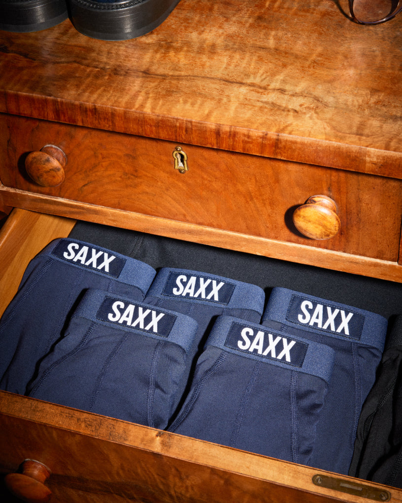 Three pairs of SAXX Boxer Briefs in white, grey, and blue, laid out neatly over a neutral background.