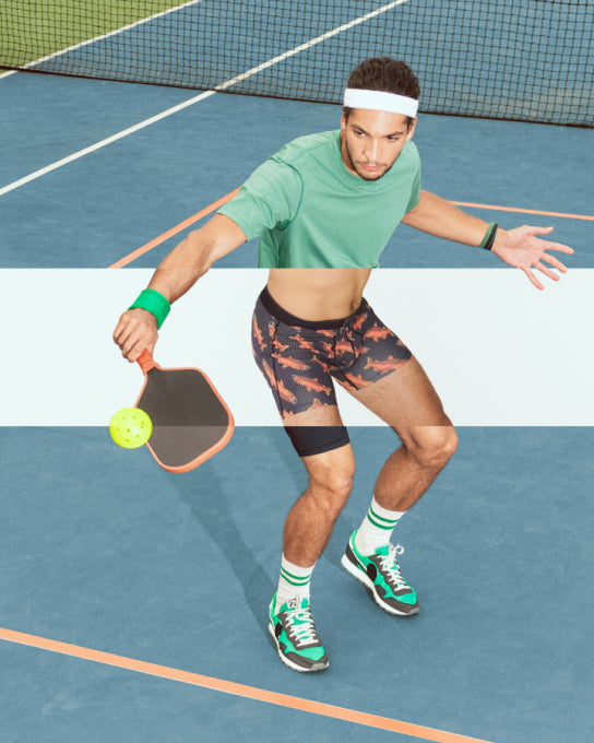 A man on a pickleball court hitting the ball with a racket, wearing a green SAXX tee shirt and black SAXX shorts. A horizontal section of the image is removed to reveal SAXX PRO performance underwear under his clothing. 