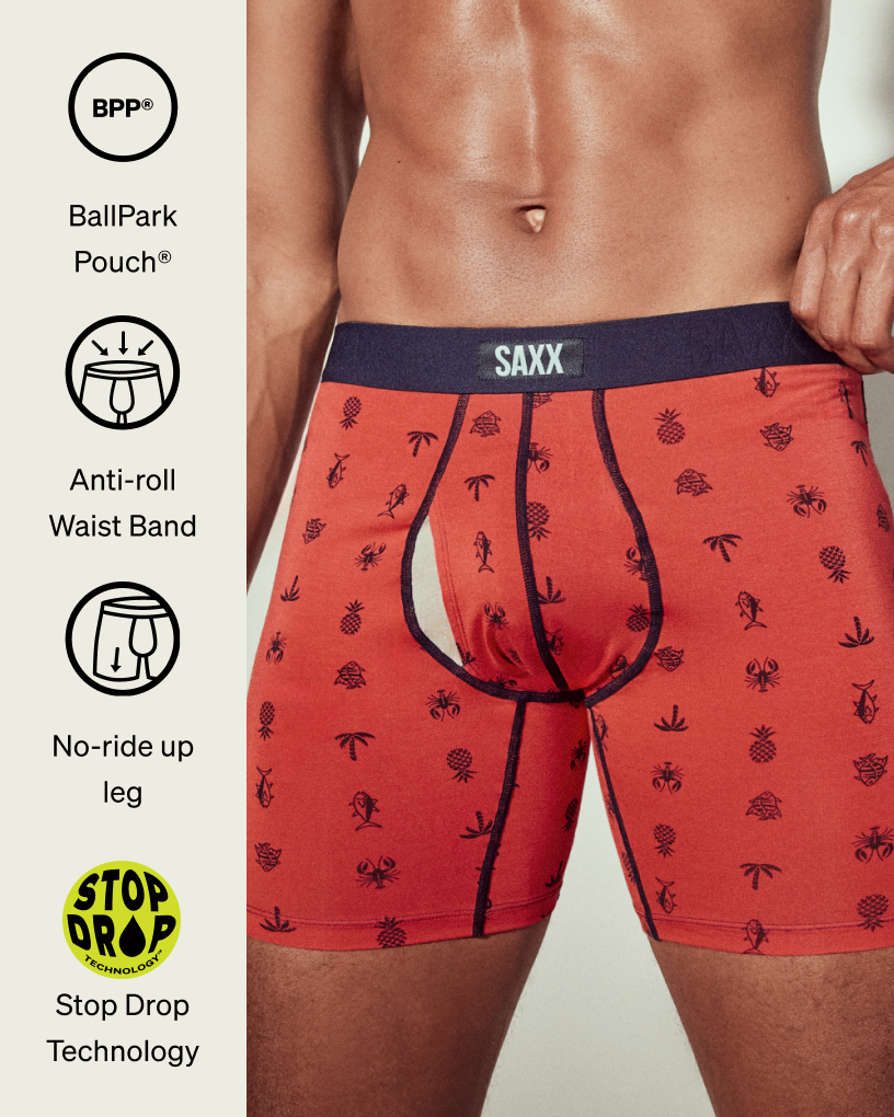 Closeup on male model wearing red patterned SAXX Vibe Xtra Boxer Briefs. With icons showing the tech features of this pair.