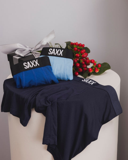 3 pairs of SAXX Boxer Briefs in shades of blue laid out over a round white table, with a wrapped gift box and holly sprig.