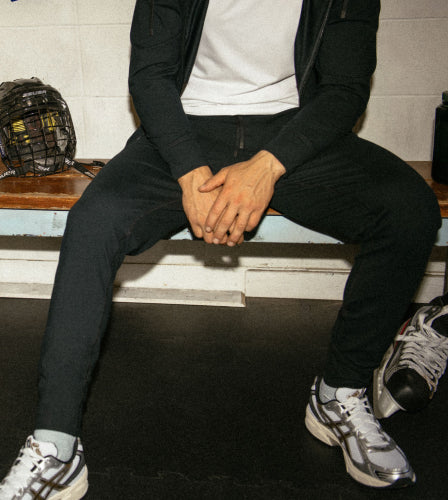 A man sitting in a sports dressing room alongside skates and a hockey helmet, wearing black SAXX joggers, a white SAXX tee, black SAXX hoodie, and running shoes. 