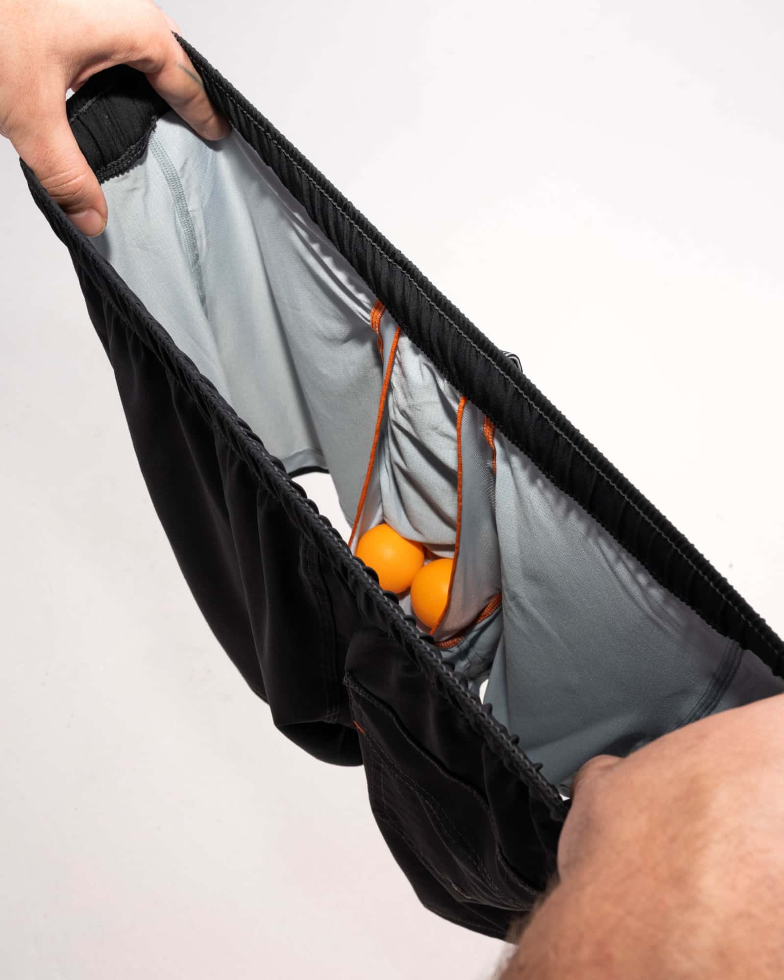 Man's hands revealing inside of 2N1 shorts showing liner with pouch holding two ping pong balls